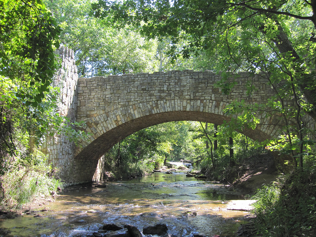 Beneath a canopy of summertime trees, a rock bridge arches over a shallow stream.