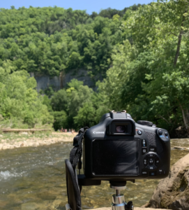 A camera is perched to photogaph a rocky stream and a towering bluff
